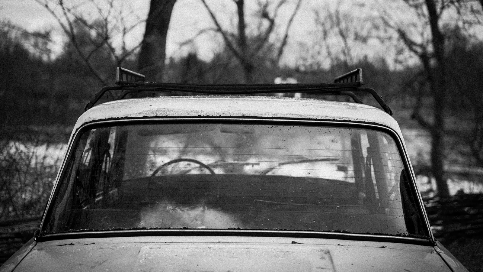 Black and white photo of an old car with a roof rack, parked among bare trees