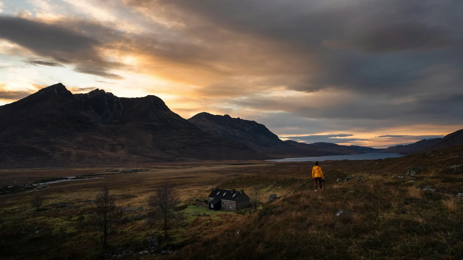 A small stone bothy and a lone walker on moorland below mountains at sunset