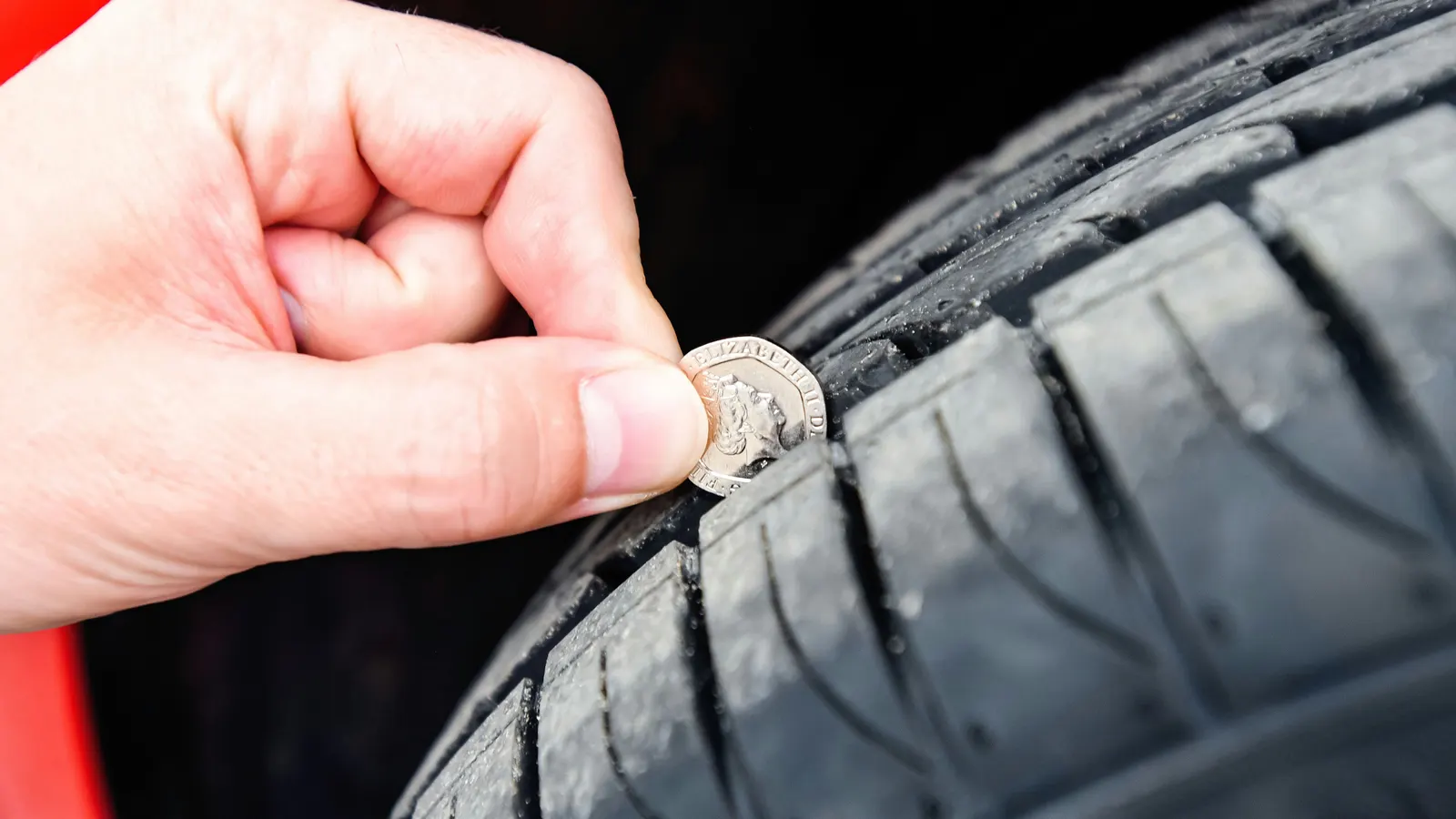 A hand holding a 20p coin in the tread of a car tyre